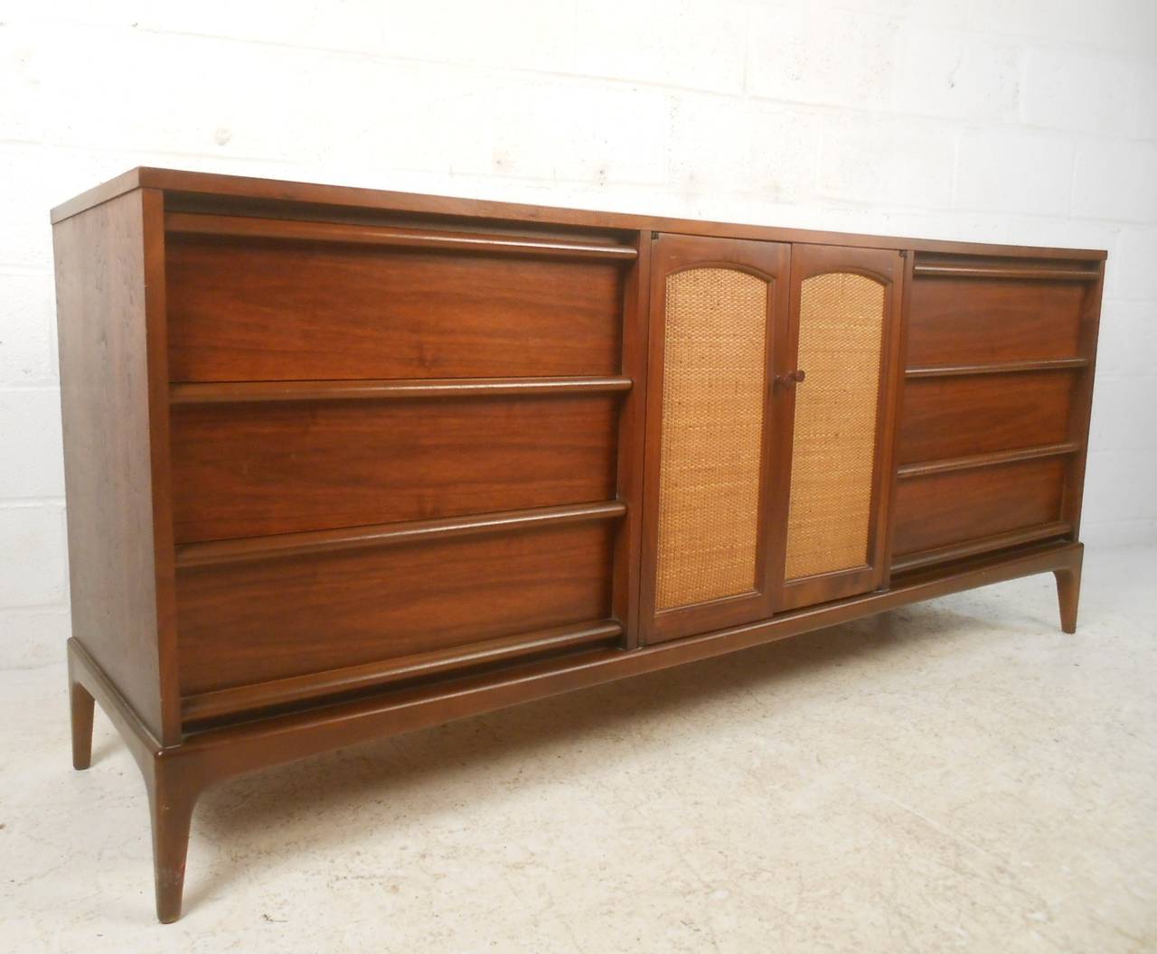 This gorgeous mid-century dresser combines modern lines with tapered legs and cane front cabinet doors to make a spectacular storage piece for any setting. With nine drawers for excessive organization the lovely wood finish of this piece contrasts 