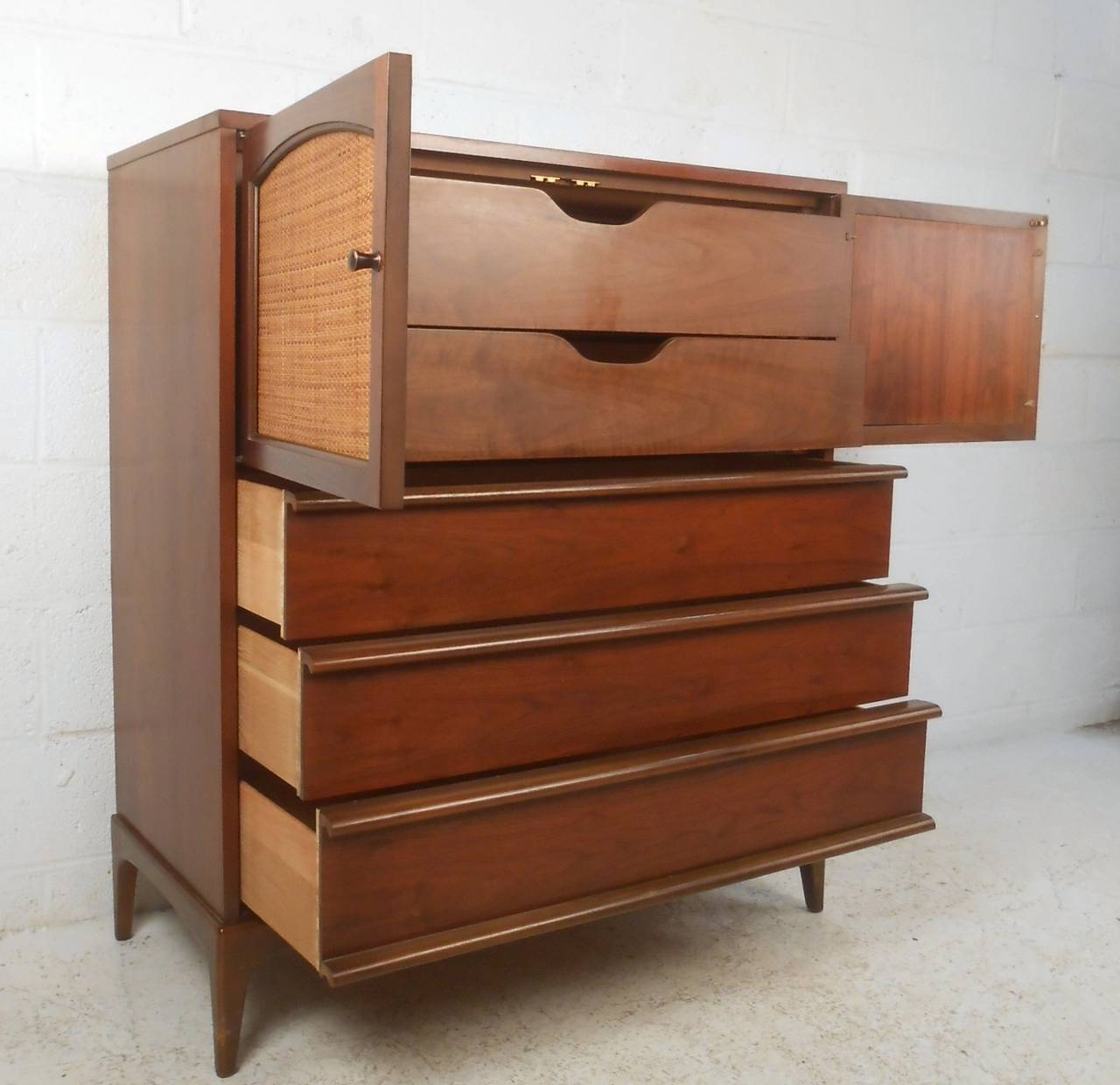 This stylish vintage highboy dresser offers five drawers for storage in any room. Wonderful walnut finish contrasts nicely with cane front upper cabinet, while carved drawer pulls and tapered legs perfectly accent it's mid-century style. Please