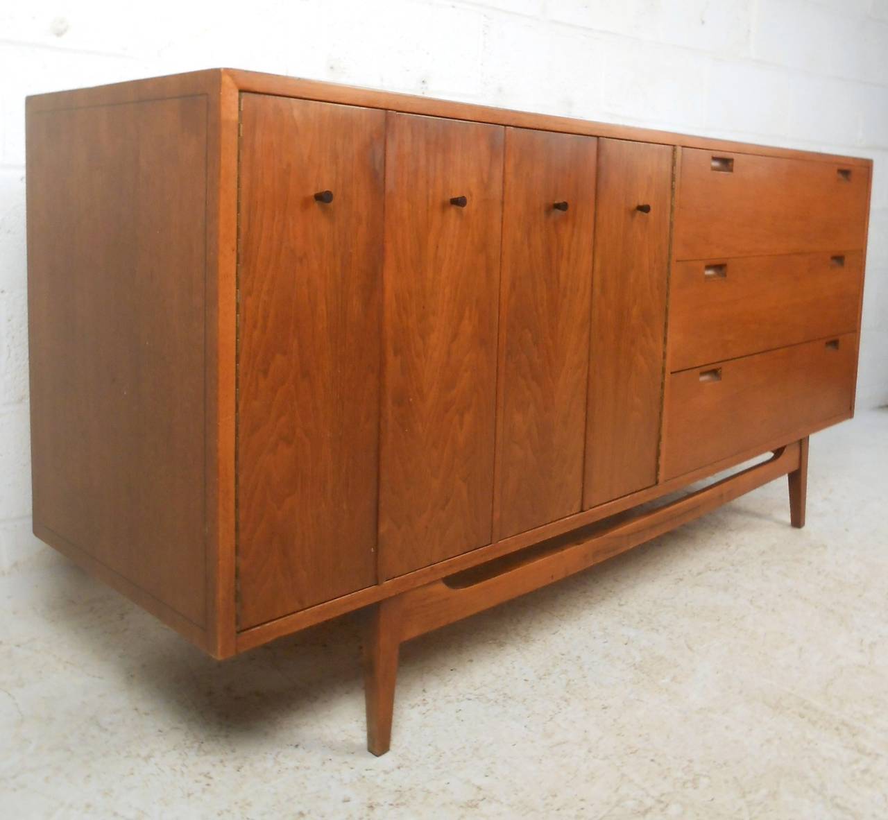 This mid-century modern dresser by American of Martinsville features unique drawer pulls, trifold door, and six spacious drawers for storage in any setting. Wonderful walnut finish and tapered legs with stretcher add to its vintage aesthetic. Please