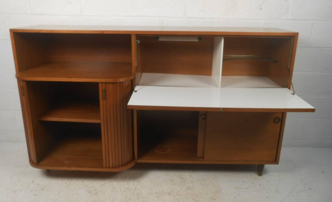 Stylish midcentury bar with tambour door storage, drop front laminate work surface with light, felt lined drawers and sliding door storage on bottom. Please confirm item location (NY or NJ) with dealer.