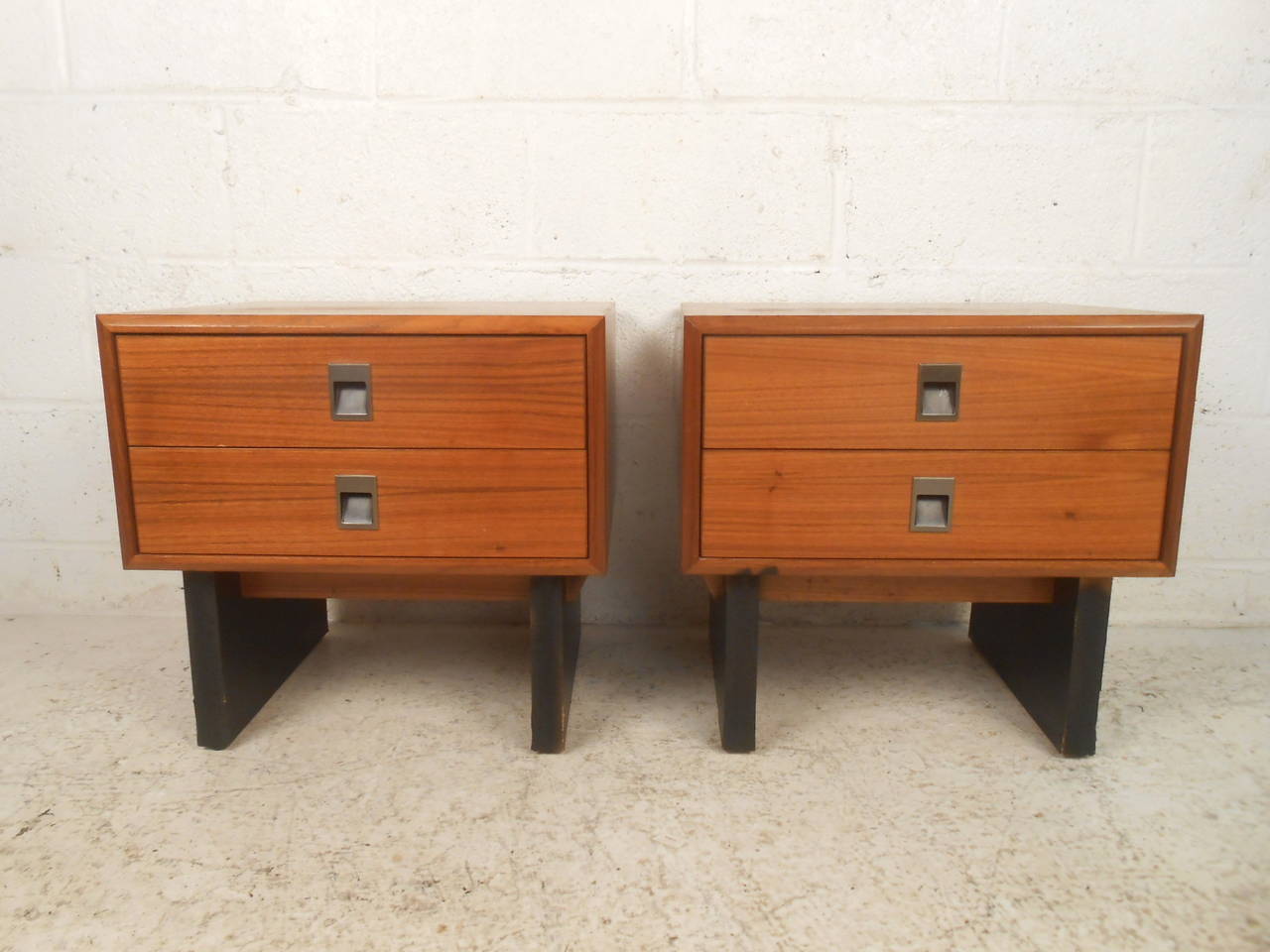 This pair of midcentury end tables feature a beautiful teak finish, two large drawers with carved pulls, and a sturdy black base which offers a modern accent and additional storage to any home or office space.

Please confirm item location (NY or