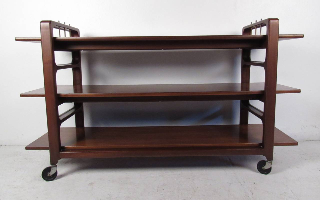 1960s American made walnut three-tiered serving cart features sculpted side supports with brass trim on top. Design by Baker Furniture Company. Please confirm item location (NY or NJ) with dealer.