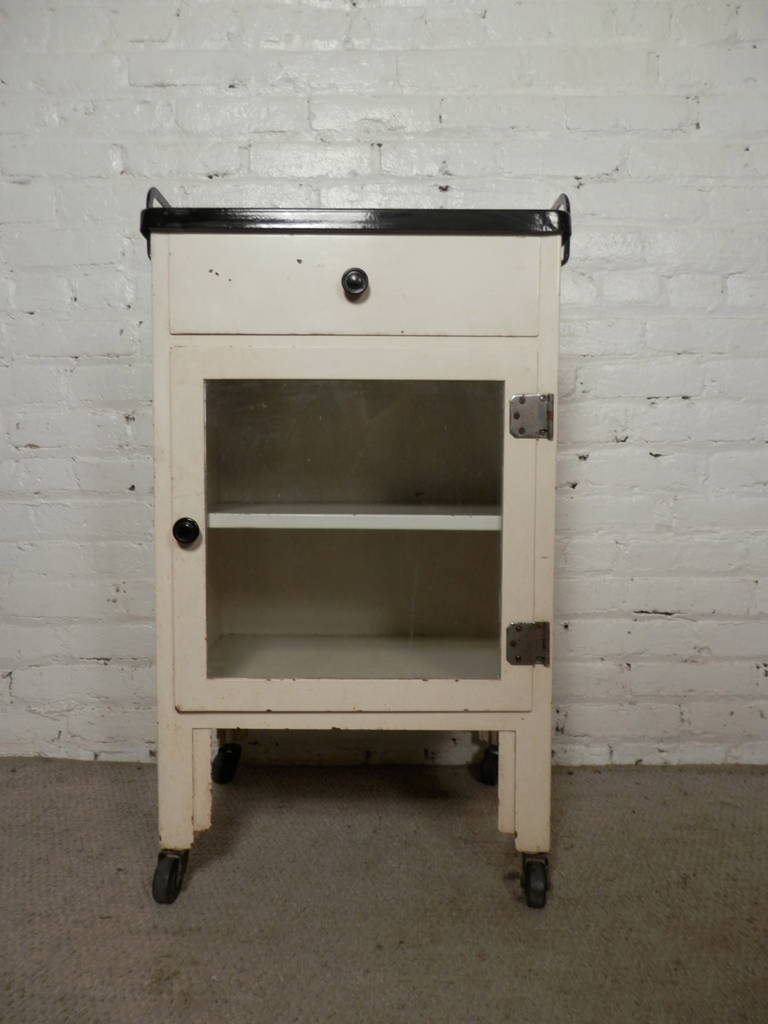 Early Industrial Style Medical Cabinet with handled top. Includes one drawer and cabinet with one shelf and glass door. All situated on sturdy metal wheeled legs. Original painted finish.

(Please confirm item location - NY or NJ - with dealer)