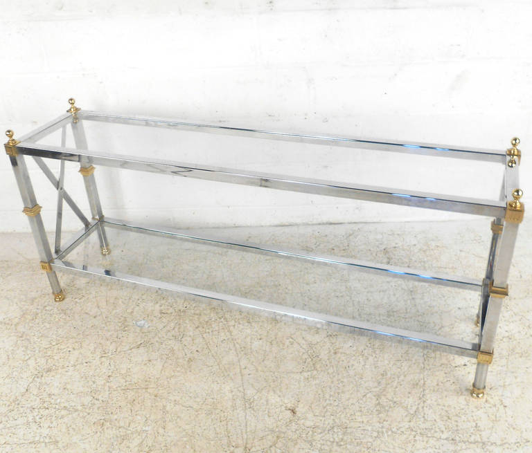 This beautiful chrome console table features beautiful brass accents and a lower shelf for secondary storage or display. A beautiful Maison Jansen style piece for use as a hall table, sofa table, or entryway display in any home or business. Please