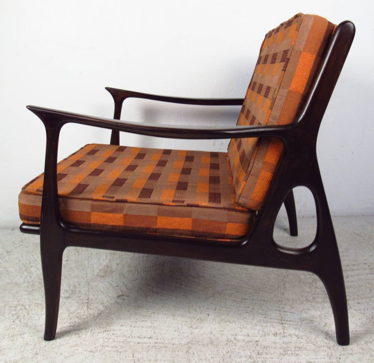 Classic Mid-Century Modern lounge chair in walnut. A sleek design with a sculpted frame, curved armrests, and a vertical slatted backrest. This attractive arm chair boasts overstuffed removable cushions covered in a plush orange upholstery. A