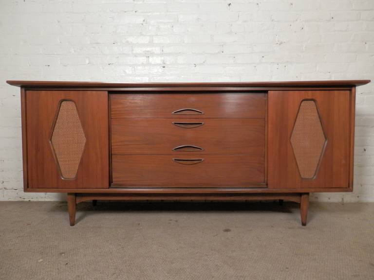 Kent Coffey made dresser with unusual design. Features beveled curved top, nine total drawers, brass detailing and sliding cane doors that hide either the side drawers or middle drawers. Interesting style that can be used as a sideboard as