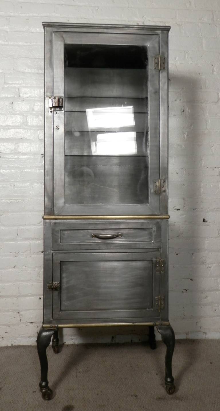 Tall metal apothecary cabinet with three clear sides of glass and three glass shelves. Unusual brass trim band around to accent the brass hardware. Working drawer and bottom cabinet. Cabinet is on casters for easy mobility. 
Works great in your