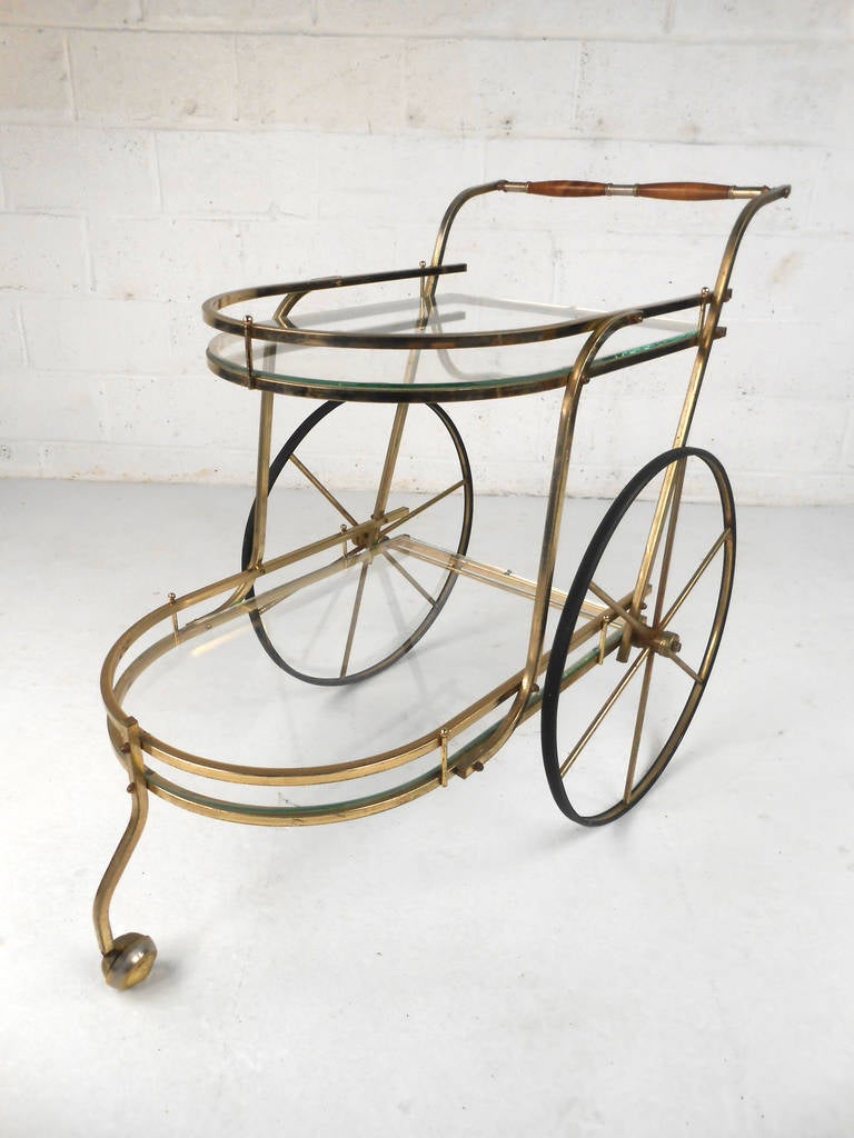 This beautiful vintage bar cart is a wonderful mix of brass and glass and makes a unique addition to any home or business for serving or display purposes. Large spoke wheels, wooden handle, and two-tier design make this a versatile serving cart.