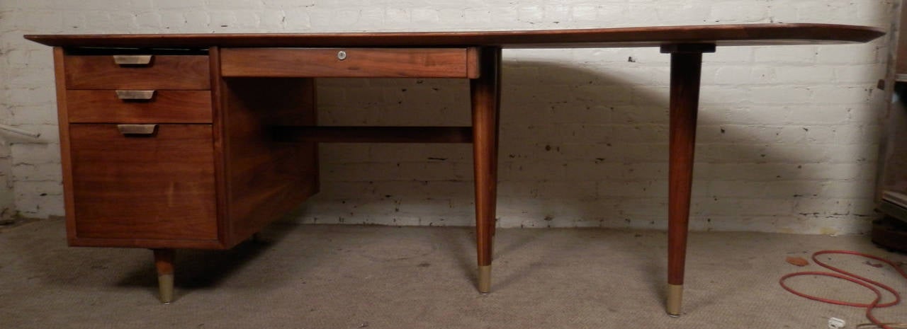 Large Mid-Century Modern American made walnut executive desk by standard.
Three drawers on the left side and one in the center all with brass handles.
Desk legs with brass caps. Unusual large kidney shape surface area.
 
(Please confirm item