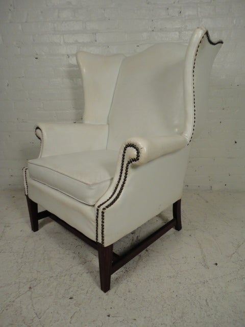 Striking white vinyl wing chair with contrasting dark wood base and brass nail heads.
