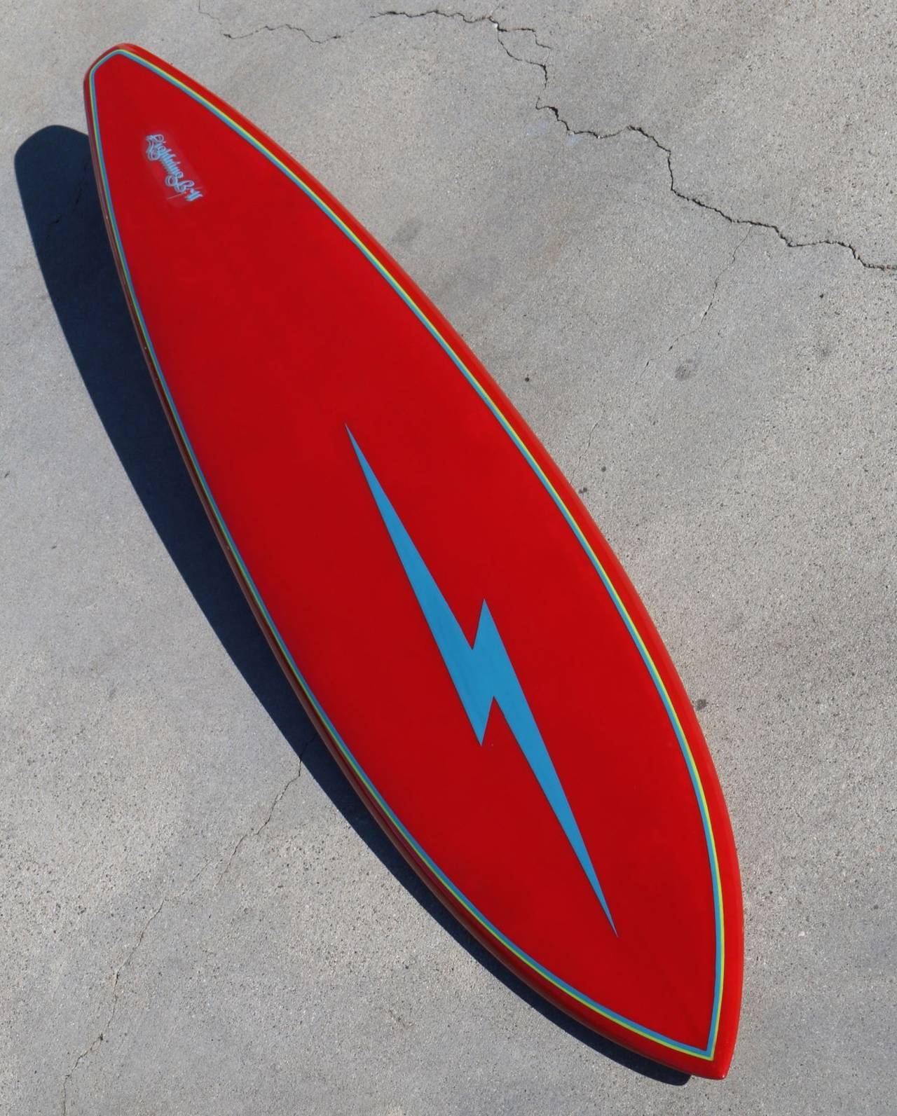 This is a fully restored 1970s surfboard with lightning bolt logo and original fin. Vibrant red deck has sky blue bolt, logo and blue and chartreuse pinstripes creating an eye-catching color combination full of verve. The bottom is the same vibrant