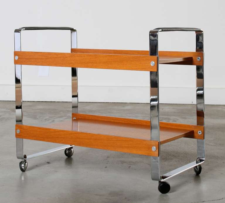 Elegant and sturdy this rolling bar cart is composed of a polished chrome frame accented with beautiful teakwood upper and lower shelves, black leather handles and chrome/black rubber articulating rolling wheels.

Stamped made in Norway on the