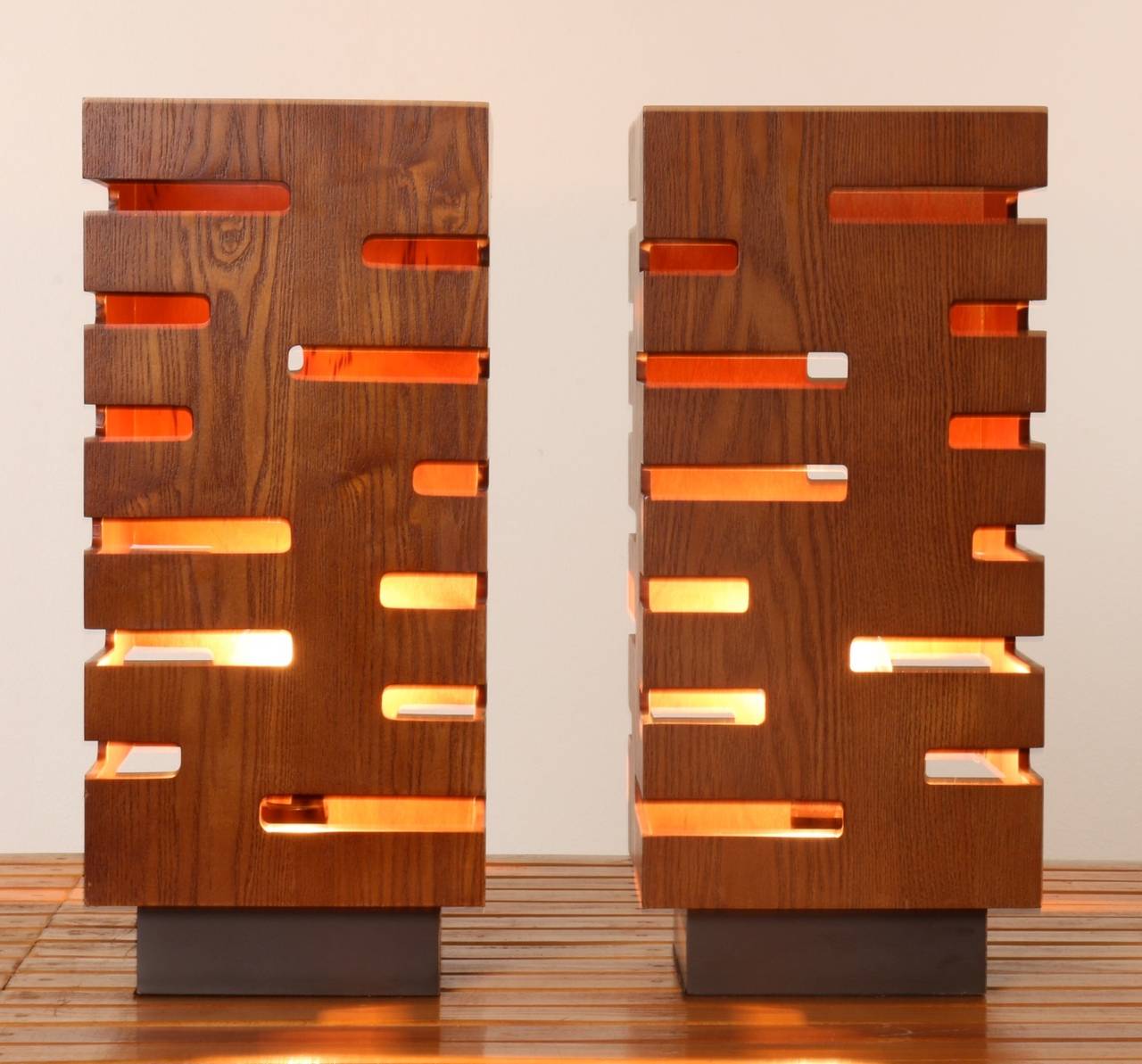 These 1970s walnut table lamps resemble rectangular towers; staggered, geometric cutouts - like open air windows create passageways from which the light shines and the air flows. Solidly built, on their original black painted wood bases, these 19