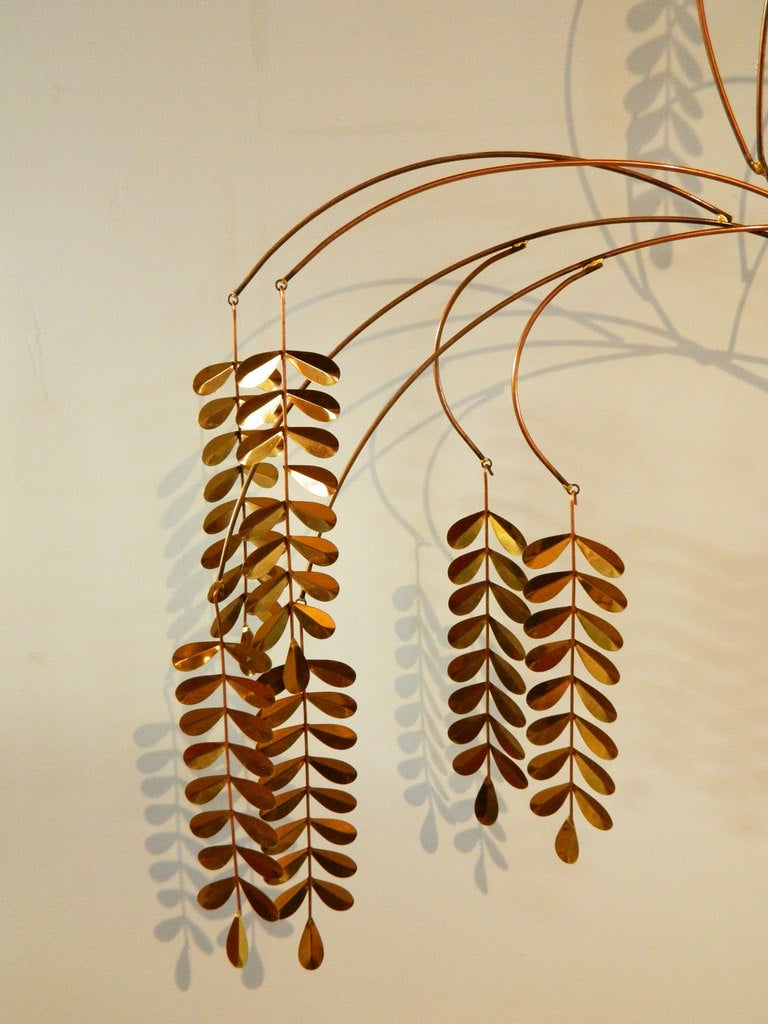 This kinetic wall sculpture has a large, free-form foundation with hanging willow branches. It is made of metal with a brass finish. Signed C Jere 1983.
