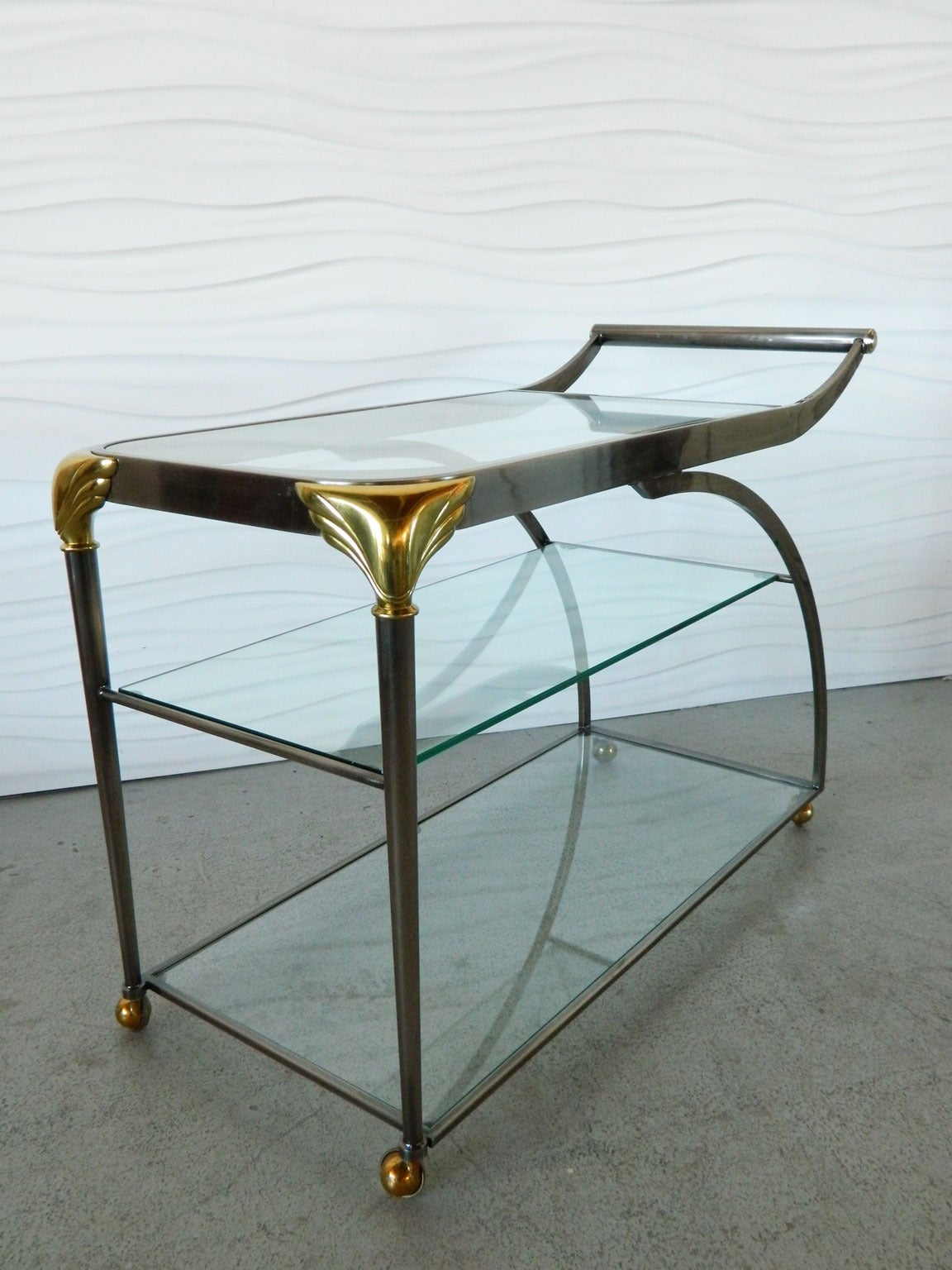 Inspired by the Deco era, this handsome bar cart with gold accets has three generously-sized glass storage/serving surfaces. The top surface has decorative etching. Casters allow the cart to be moved easily.