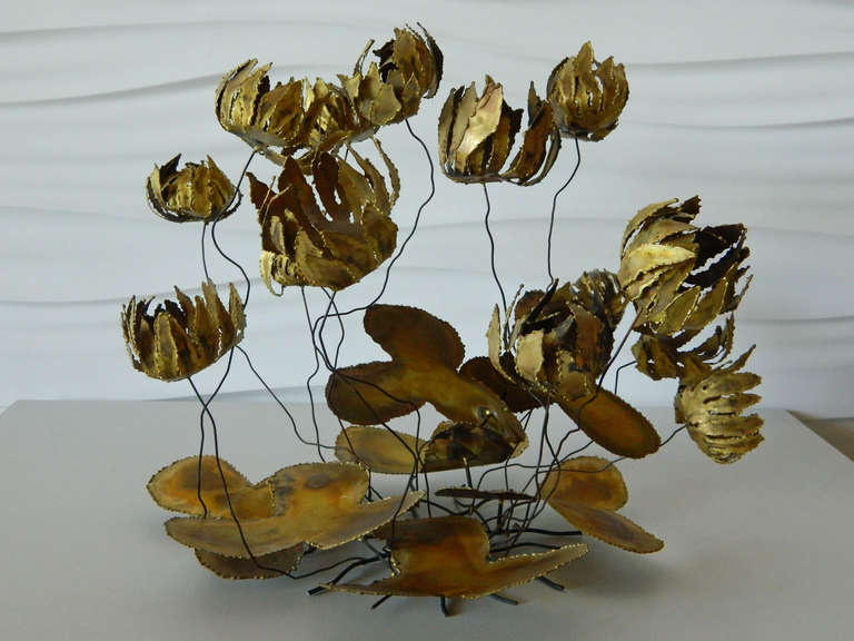 Delicate metal flowers suspended by thin wires hover above torch-cut lily pads to create a table-top kinetic sculpture in the style of Curtis Jere.
