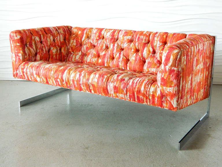 This tufted loveseat appears to float from a chrome, cantilevered base. It is covered in a linen-like fabric with a bright, watercolor abstract.