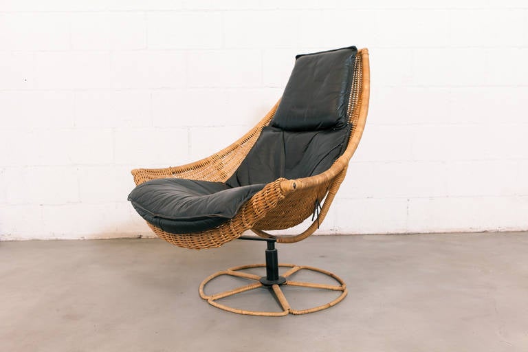 Woven rattan frame swivel lounge chair with bat-like armrests and scooped seat. Black leather cushion and headrest with great patina. Enameled black metal swivel support and rattan wrapped base. Original condition with some breakage or wear to