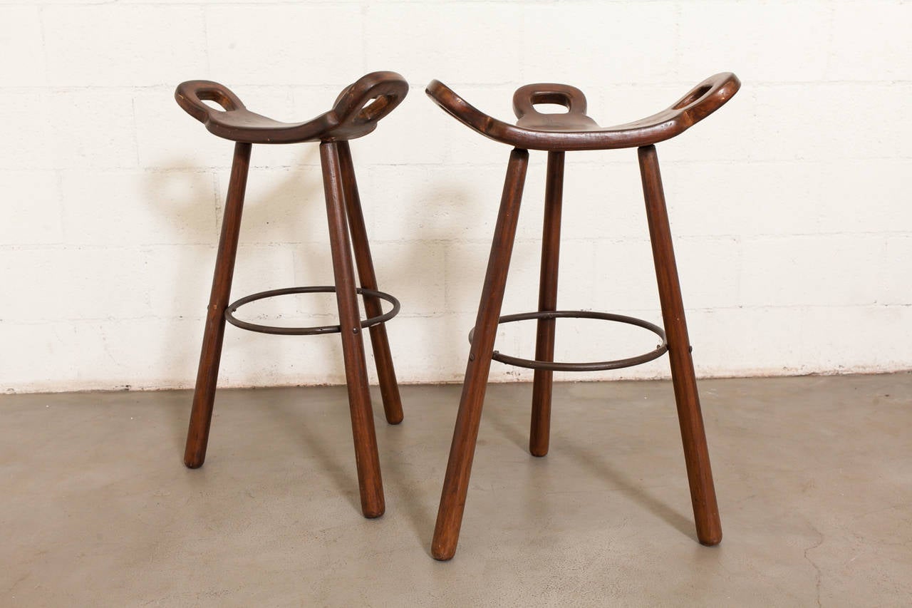 Elegantly Carved Triopod Bar Stool Seat with Looped Detail and Iron Foot Rest. Some cracking to the wood. Original Condition. Set Price