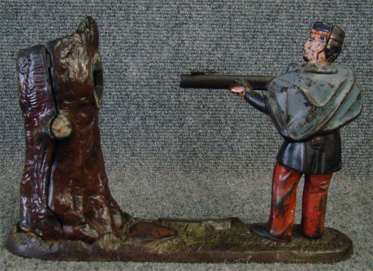 “Creedmoor” mechanical bank with original polychrome decoration. The soldier is wearing a blue cape, blue jacket, red pants and a blue hat. The paint is worn, as expected from the bank’s age and usage. The large rock reads 