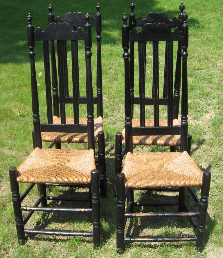 Set of four banister-back side chairs in old black paint. The chairs boast beautiful finials and crests and natural rush seats in excellent condition. The nice height of the chair backs makes for an impressive presence around a table. The chairs