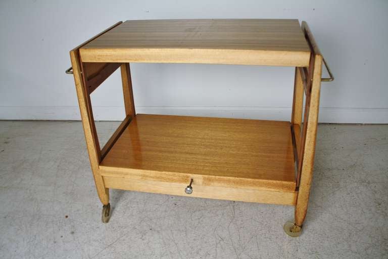 1950s two-tiered tea cart in sycamore wood with a mechanism that allows the bottom shelf to rise and convert to a server.  Designed by John Keal for Brown and Saltman.  On casters with brass handles on each side.  Measurements when expanded are 31