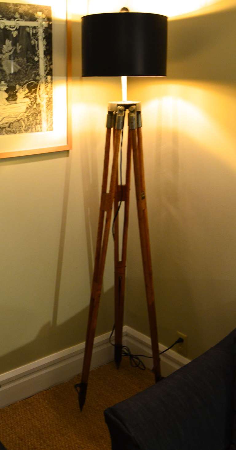 Industrial strength wooden surveyor tripod as floor lamp. Standing atop steel ground spikes, solid oak legs lead to galvanized steel platform that has been fitted with 3-way light socket, 8