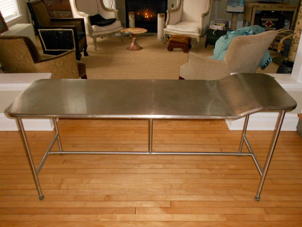 Mid-century stainless steel medical exam table with head bump. The form of this table is exquisitely fluid. Its minimalist lines creating a wonderful tension between the rigid, linear legs and crosspiece, and the curved, liquid flow of the stainless