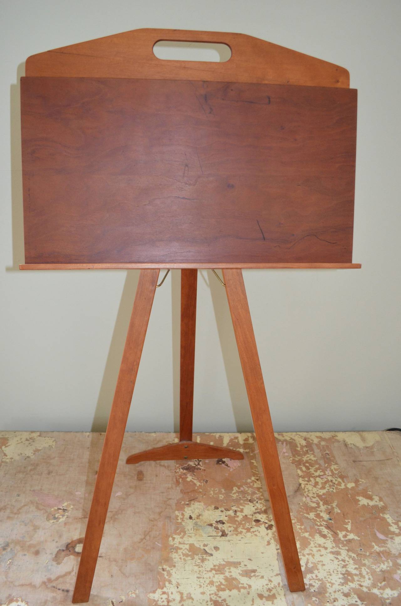 Mid-century display easel on a collapsible stand with storage compartment. Simplicity personified constructed in what appears to be cherrywood. The back leg of the stand hinges out and locks in place for display purposes. Folds in for slim-lined