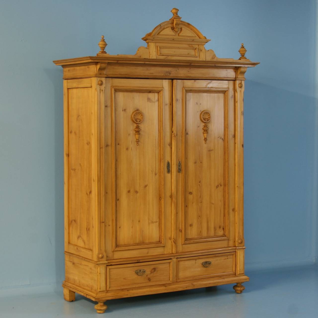 Impressive in stature, this pine armoire has a strong visual presence due to the turned finials and decorative upper crown. Even the paneled doors have decorative carved elements, adding a sophisticated touch to the piece. It is a 2-door over