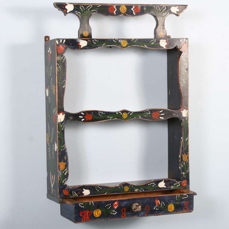The vibrant painted flowers (tulips) and colors of this shelf are all original. The drawer functions easily and is dated 1898. This delightful piece may hang in a hallway, bathroom, or virtually any space.