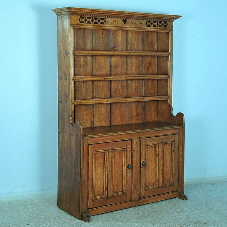 Lattice carved detail and the date of 1824 grace the top of this charming cupboard from England. The pine has aged gracefully and reflects a lovely warm patina. The wood feet extend forward slightly from the front, door pulls are metal and stoppers