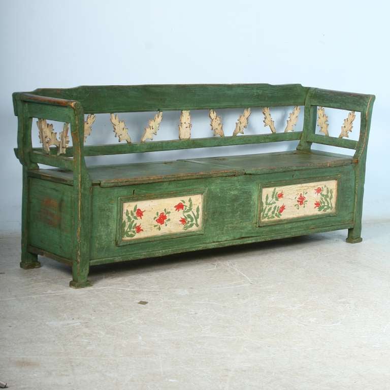 The paint is all original on this green bench with floral details. The carved details along the back and curved arms were a traditional style of the period. This bench has 2 separate hinged seats, revealing storage below.