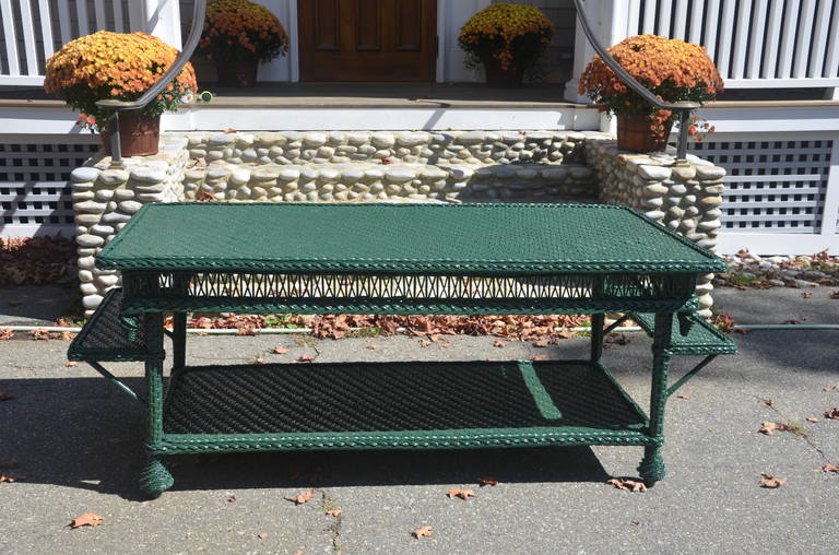 Antique wicker library table in green paint. All shelves are woven in a herringbone pattern with reverse finials on the four corners of the top shelf. This is a substantial, heavy piece.

Table overall length is 82