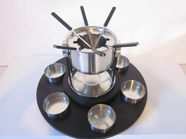 A stainless steel Fondue pot set with cooking pot,inserts topper for forks,pot stand,bottom plate,six condiment dishes,six color coded forks and all sitting on a  ebony wood toned rotary 