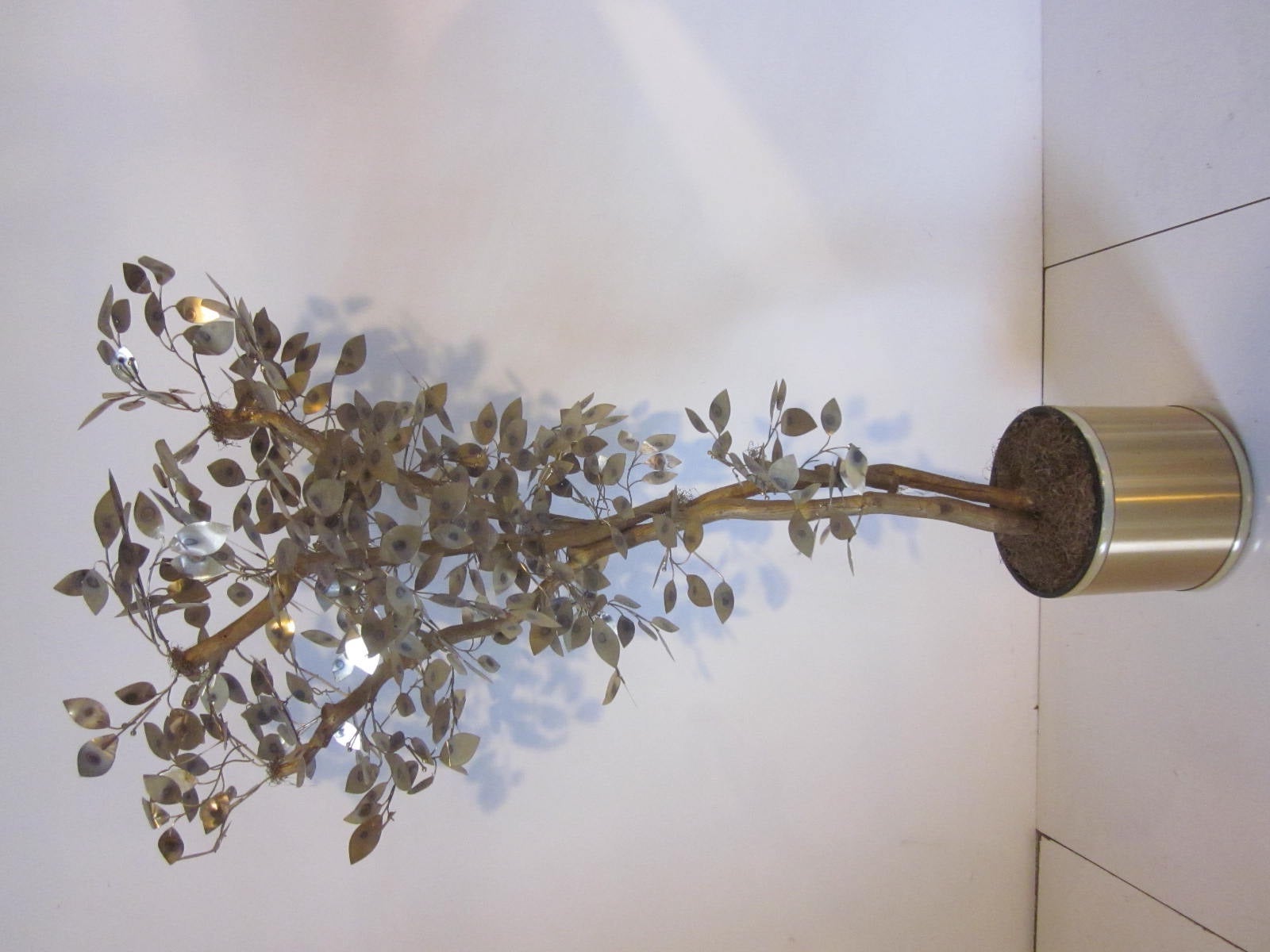 A Jere tree sculpture with brass metal flamed patinated leaves and stems mounted on a gilded natural wooden tree trunk sitting in a brushed brass planter with plastic insert.