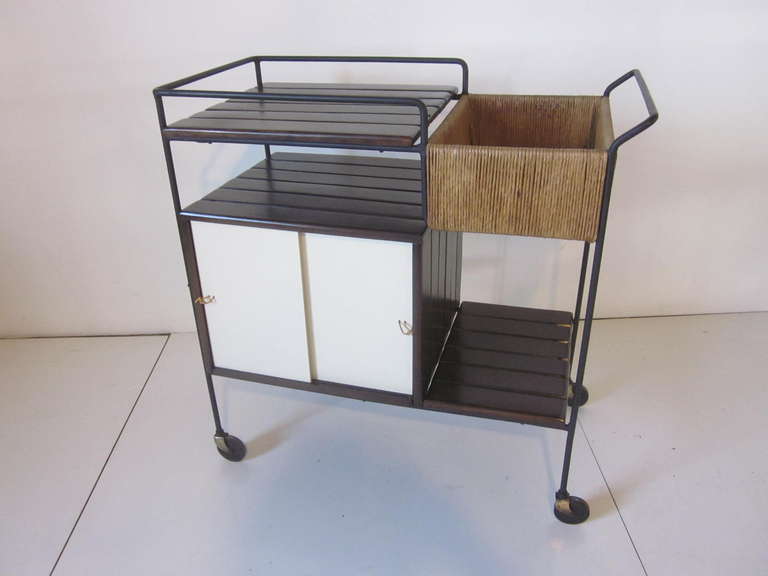 A Umanoff rolling bar cart with lower storage and sliding doors on both sides, bottle , glass holder and serving area all sitting on a black iron frame. A  raffia  wrapped area and the wood has been restored in a dark and rich ebony tone made in