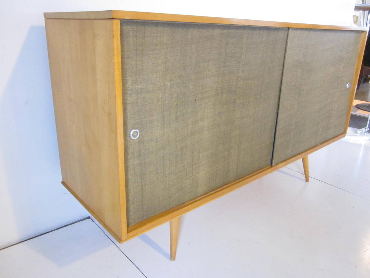 A very early McCobb light maplewood credenza with grass cloth covered cement doors and aluminum pulls, four internal drawers and a adjustable shelve sitting on a matching platform base with conical legs. From the Planner Group series manufactured by