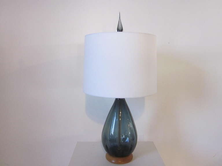 A nice mid sized hand-blown Blenko table lamp with the original matching finial sitting on a maple wood base and off white linen shade. The glass is a blue gray and retains the original manufactures stamp to the bottom felt .