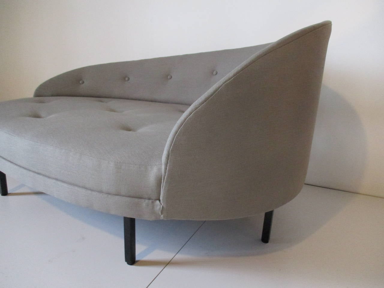 A sculptural chaise longue chair upholstered in a gray, taupe toned linen with satin black boxed metal legs, button tufted back and bottom.
