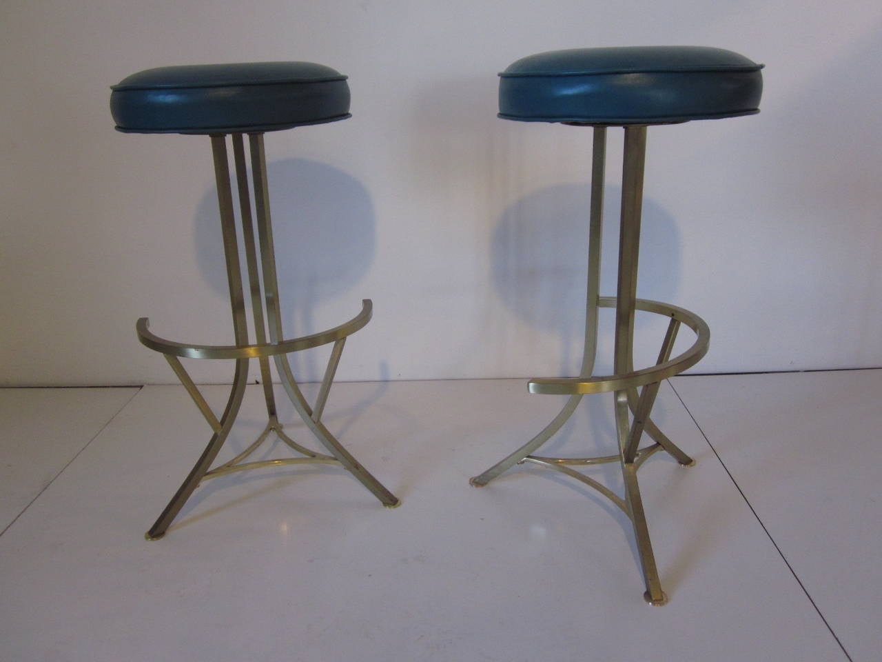A pair of solid brass bar stools with half moon crescent shaped foot rests, brass foot pads and upholstered swiveling round cushion with big brass button . In a dark teal toned turquoise leatherette with the cushion dia. 13.50
