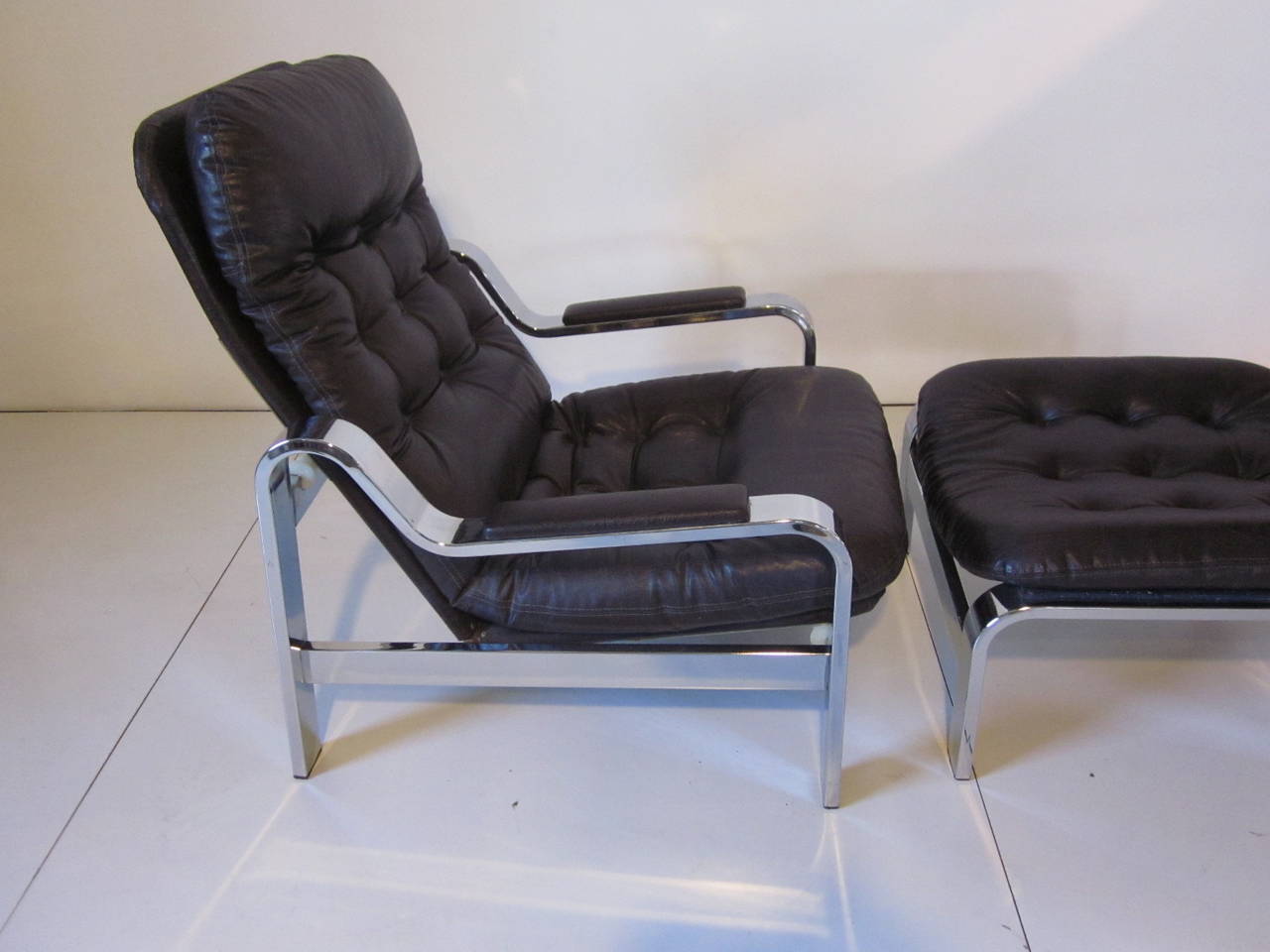 A chromed framed lounge chair with rolling, sliding recliner mechanism and matching ottoman both upholstered in a textured leatherette with button tufting. Heavy and well made, by the Selig Furniture company, ottoman measurements 15