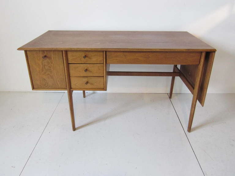 A Mid Century Walnut drop leaf desk with center pencil drawer,three smaller drawers and a file. It has matching wooden pulls with bronze toned inserts and a finished desk back.The fold down leaf extends the desk to a total of 71.75