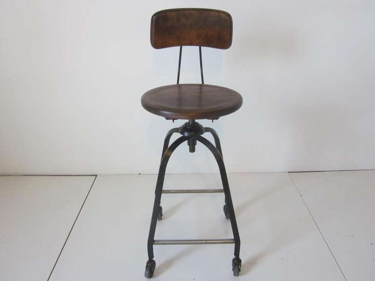 A tall Industrial stool with wood seat bottom and back, metal frame on wheels with corkscrew adjustable seat height. Has some painted details to the screws and has a stainless foot rest area.