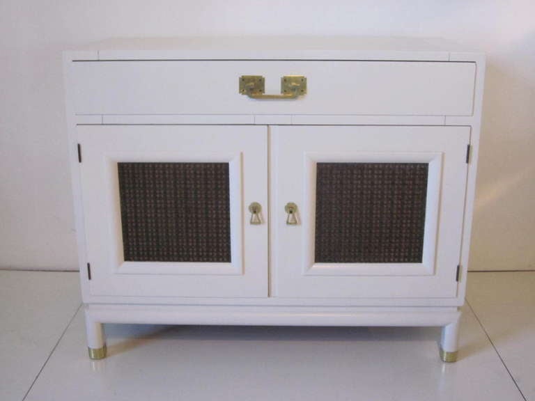 A bone satin white two door Rutili cabinet with brass hardware and foot caps,metal woven door panel inserts and top drawer with dividers. Inside it has an adjustable shelve,manufactured by the Johnson Brothers Furniture Company,Grand Rapids,Mich.  