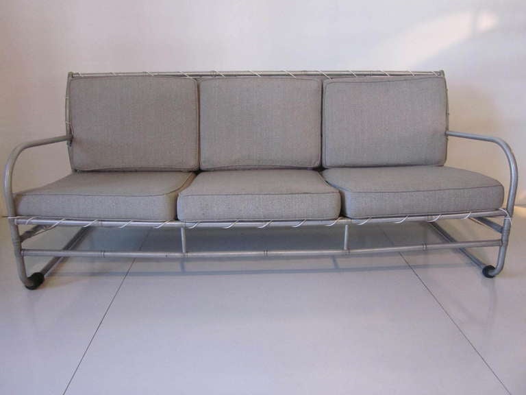 A rare McArthur three cushion sofa with aluminum tubular frame, black hockey puck feet and upholstered in a nice gray toned sail cloth type fabric. This rare sofa has a great architectural tubed frame design with complex bends and fittings from the