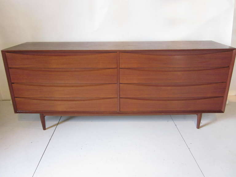 A Vodder eight drawer teakwood dresser with cut out upper pulls sitting on a simple base with conical teak legs. The upper right side drawer has dividers and a sliding tray for jewelry, watches, wallet etc. Finished on the backside with the same