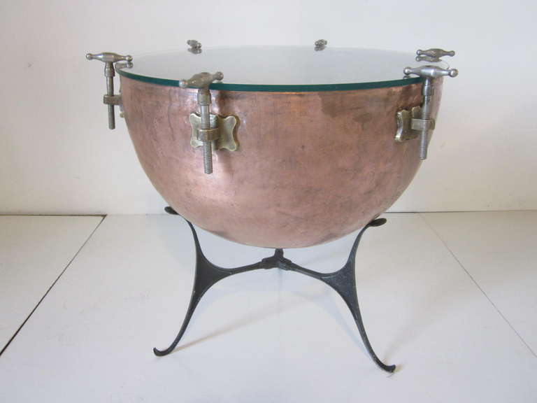A vintage copper Kettle drum that has been repurposed as a side table with glass top, brass fittings and steel tuning screws. The table sits on a folding black metal support.