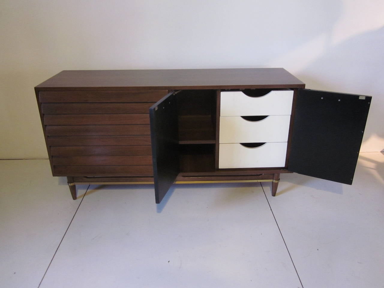 A dark walnut credenza with three side drawers, two black doors, inside three off-white small drawers and a storage area complemented by brass pulls and detailed trim to the lower stretchers.