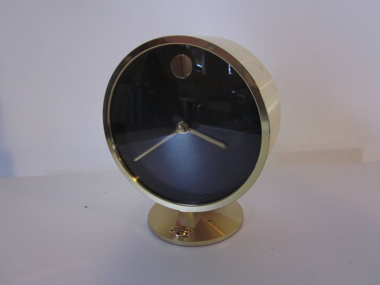 A Howard Miller table clock in a satin brushed brass finish with black face and 12 o'clock positioned brass dot and hands. It has a glass covered face, pedestal base with small logo from a company used as a presentation piece. Pull off back revels